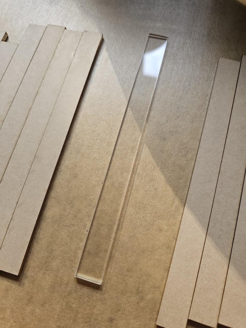 May include: A clear, rectangular acrylic strip is positioned diagonally, surrounded by several light brown, rectangular wooden strips. The acrylic strip reflects light, contrasting with the matte finish of the wood. The items are arranged on a neutral-toned surface.