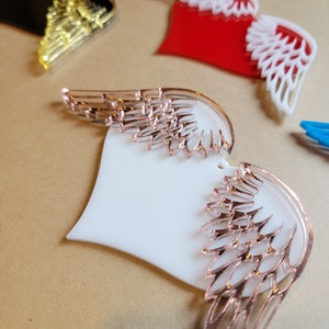 May include: A white heart-shaped ornament with rose gold wings. The wings are detailed with a delicate, intricate design.