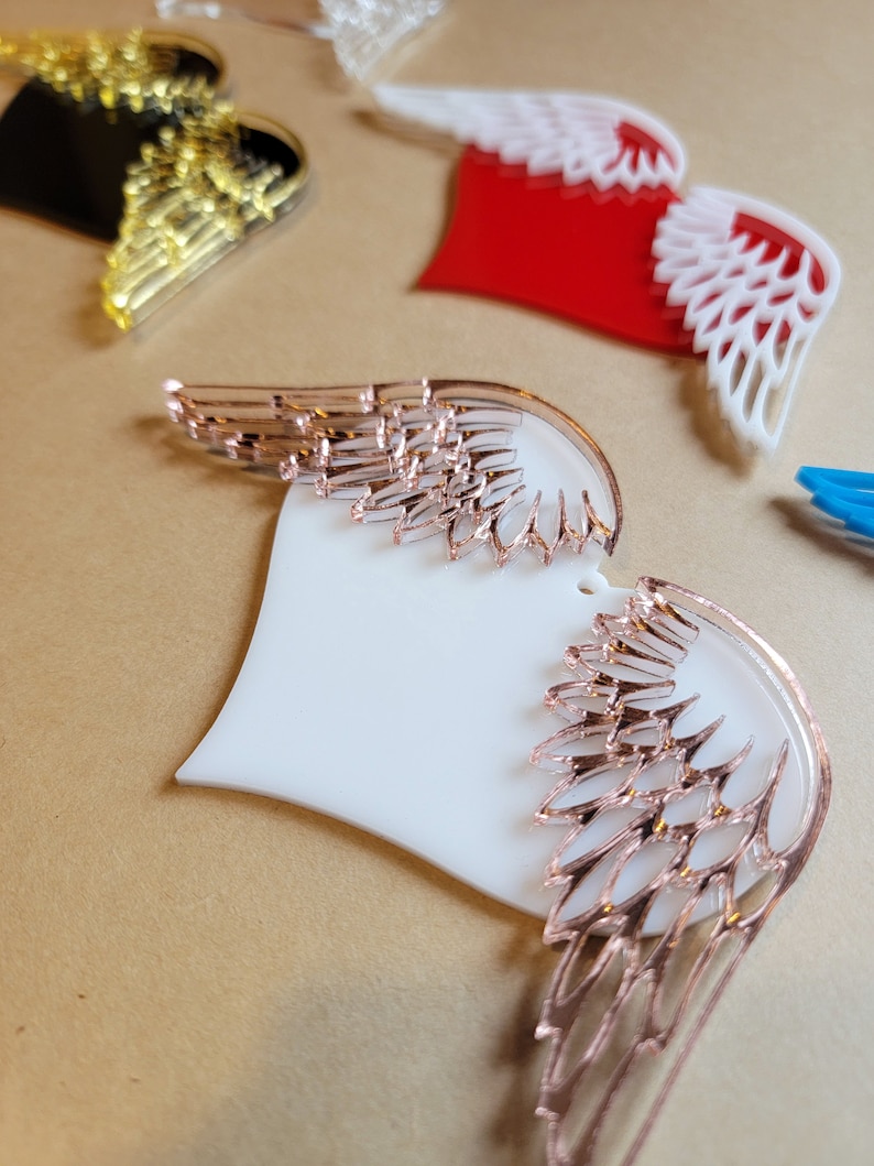 May include: A white heart-shaped ornament with rose gold wings. The wings are detailed with a delicate, intricate design.