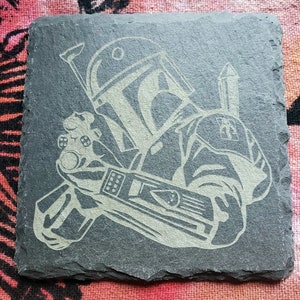 Pu&ograve; includere: Un sottobicchiere in ardesia nera con un'immagine incisa in bianco del personaggio di Star Wars, Boba Fett, che tiene un fucile blaster. L'immagine &egrave; dettagliata e mostra l'elmetto, l'armatura e l'arma di Boba Fett.