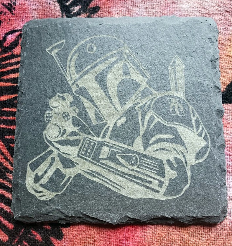 Pu&ograve; includere: Un sottobicchiere in ardesia nera con un'immagine incisa in bianco del personaggio di Star Wars, Boba Fett, che tiene un fucile blaster. L'immagine &egrave; dettagliata e mostra l'elmetto, l'armatura e l'arma di Boba Fett.
