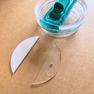 May include: A clear plastic lid with a teal green slider. The lid has a white silicone ring and a clear plastic ring. There are two separate clear plastic pieces, one with a hole in the centre.