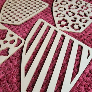 May include: Five different wooden cutouts with various patterns. The cutouts are in the shape of a shield and have a light brown color. The patterns include stripes, polka dots, a checkered pattern, and a leopard print.