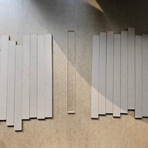 May include: Arrangement of rectangular, light-colored wooden pieces, some with varying heights, alongside a single, clear, rectangular piece. The pieces are arranged on a neutral-toned surface, creating a minimalist aesthetic.