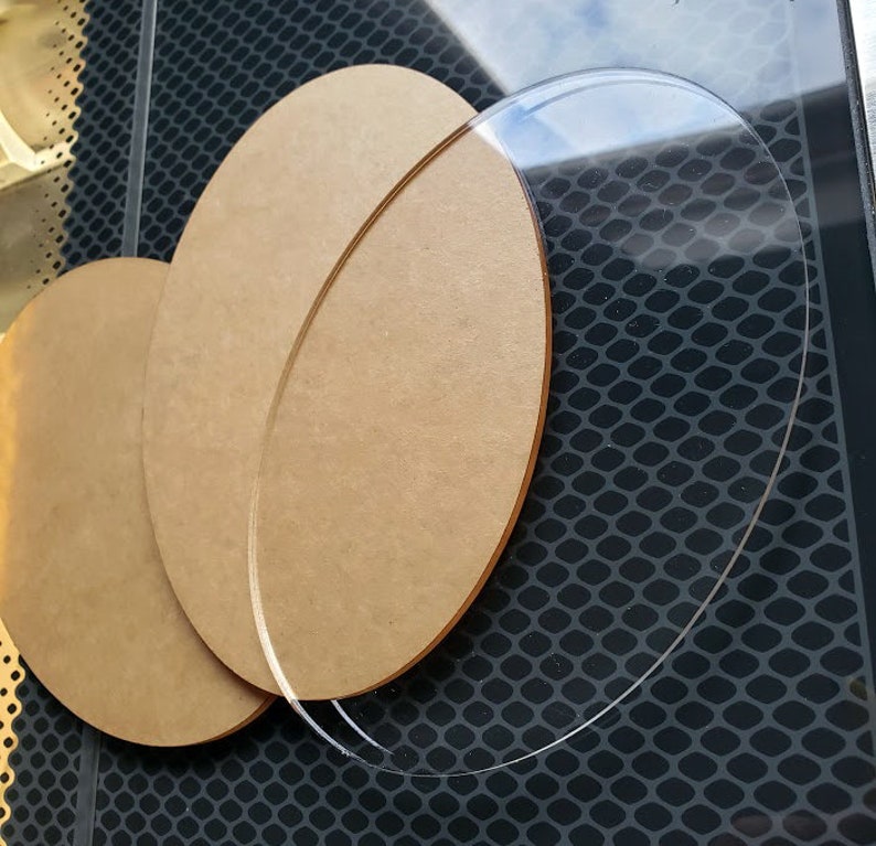 May include: Two round, wooden craft blanks, one larger than the other, are stacked on top of a clear, round acrylic blank. The blanks are all on a black, textured surface.