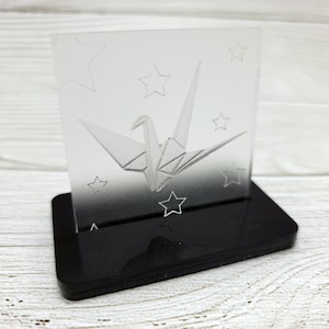 May include: A clear acrylic paperweight with a 3D etched design of a paper crane and stars. The paperweight sits on a black acrylic base.
