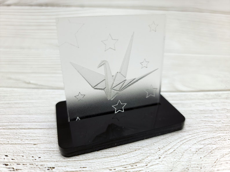 May include: A clear acrylic paperweight with a 3D etched design of a paper crane and stars. The paperweight sits on a black acrylic base.