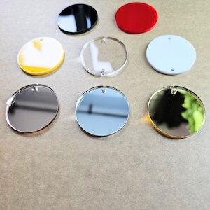 May include: Assortment of eight round acrylic blanks in various colors, including black, red, white, yellow, clear, silver, and gold. The blanks have a hole at the top for hanging.
