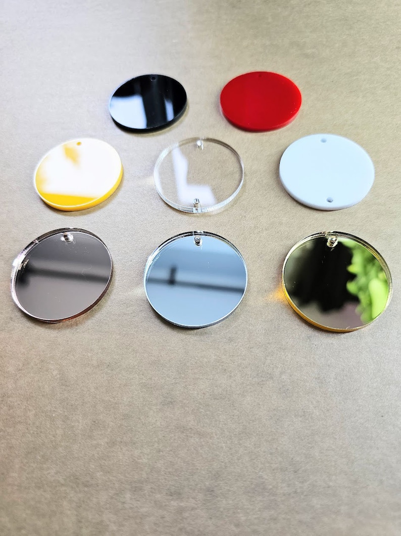 May include: Assortment of eight round acrylic blanks in various colors, including black, red, white, yellow, clear, silver, and gold. The blanks have a hole at the top for hanging.