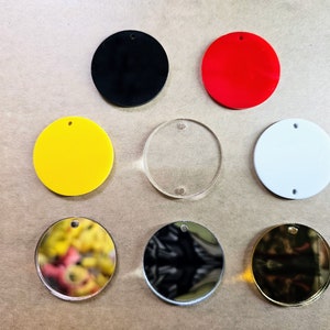 May include: Assortment of eight round blank acrylic ornaments with a hole at the top for hanging. The ornaments are in various colors, including black, red, yellow, white, clear, gold, and silver.