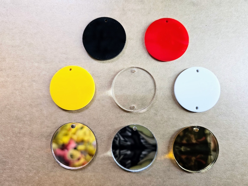 May include: Assortment of eight round blank acrylic ornaments with a hole at the top for hanging. The ornaments are in various colors, including black, red, yellow, white, clear, gold, and silver.