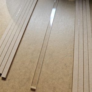 May include: An overhead shot shows several thin, rectangular pieces. There are two stacks of cardboard strips and one clear, rectangular plastic strip. The items are arranged on a light brown surface.