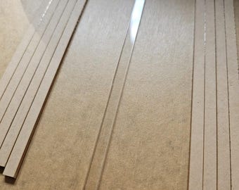 May include: An overhead shot shows several thin, rectangular pieces. There are two stacks of cardboard strips and one clear, rectangular plastic strip. The items are arranged on a light brown surface.