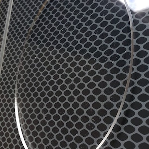May include: A clear, round, acrylic disc with a slightly textured surface. The disc is placed on top of a black, patterned surface.