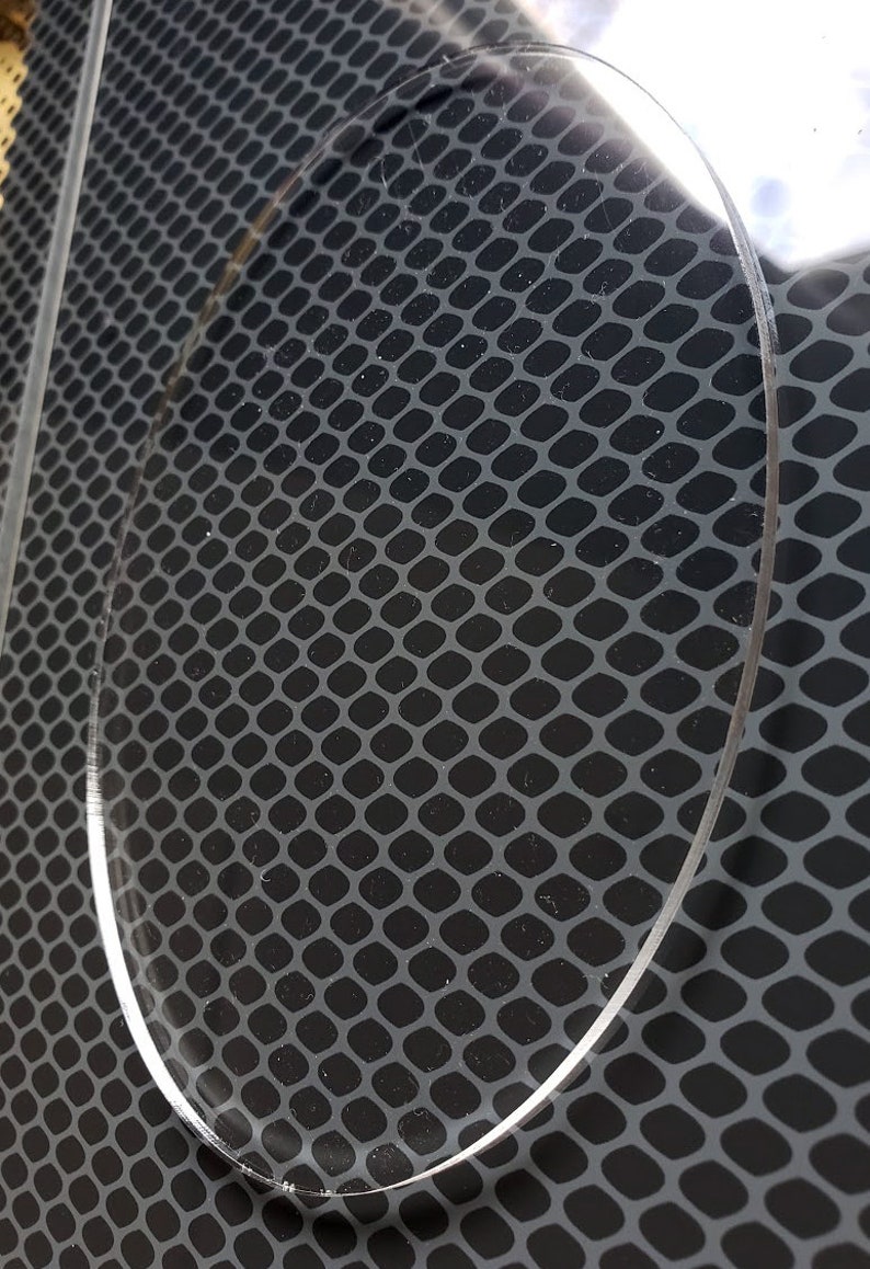 May include: A clear, round, acrylic disc with a slightly textured surface. The disc is placed on top of a black, patterned surface.
