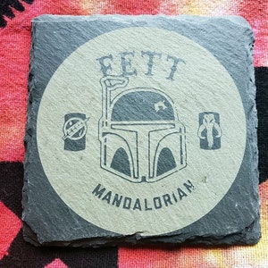 Pu&ograve; includere: Un sottobicchiere in ardesia grigia con un cerchio bianco che presenta un'illustrazione in bianco e nero di un elmetto mandaloriano con il testo "FETT MANDALORIAN".