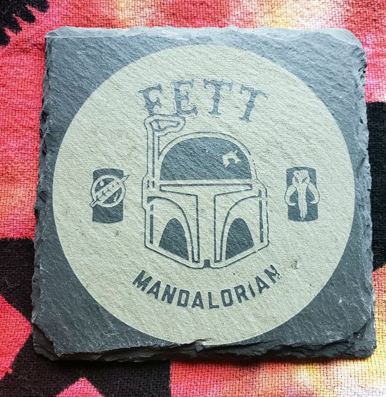 Pu&ograve; includere: Un sottobicchiere in ardesia grigia con un cerchio bianco che presenta un'illustrazione in bianco e nero di un elmetto mandaloriano con il testo "FETT MANDALORIAN".