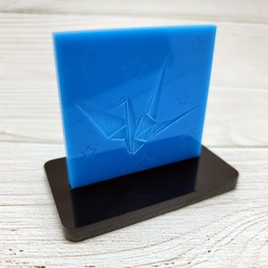 May include: A blue acrylic plaque with a white origami crane design and stars. The plaque is on a black stand.