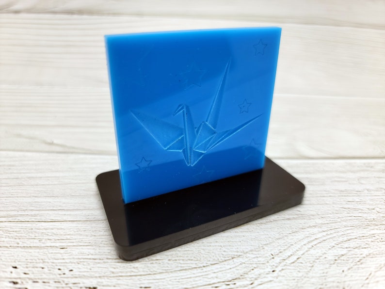 May include: A blue acrylic plaque with a white origami crane design and stars. The plaque is on a black stand.
