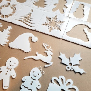 Peut inclure: Un ensemble de 14 pochoirs en plastique blanc représentant divers motifs sur le thème de Noël, notamment un renne, un bonhomme de neige, un personnage en pain d'épices, un sapin de Noël, un flocon de neige, une feuille de houx, une couronne, un coffret cadeau et un bonnet de Père Noël.
