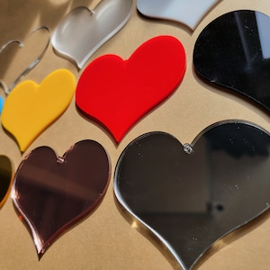 May include: Assortment of heart-shaped acrylic blanks in various colors, including red, yellow, blue, black, gold, and silver. The hearts are all approximately the same size and have a smooth, glossy finish.