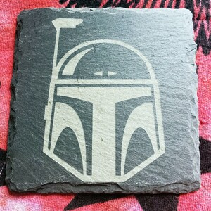 Pu&ograve; includere: Un sottobicchiere in ardesia nera con una silhouette bianca di un casco di Boba Fett di Star Wars.