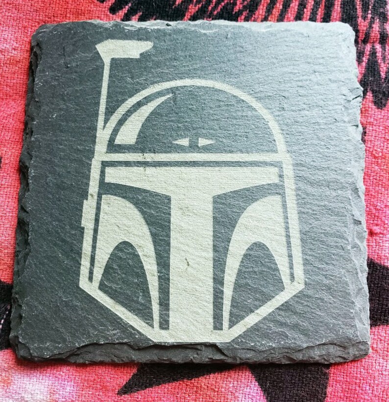 Pu&ograve; includere: Un sottobicchiere in ardesia nera con una silhouette bianca di un casco di Boba Fett di Star Wars.
