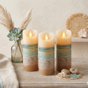 May include: Three battery-operated pillar candles with a beach design. The candles have a cream top, a blue and tan stripe, and a sandy base. They emit a warm, flickering light, creating a relaxing ambiance.