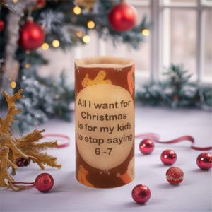May include: A cylindrical Christmas candle with a brown background and the text "All I want for Christmas is for my kids to stop saying 6-7." The candle is surrounded by red Christmas ornaments.