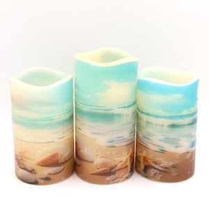 May include: Three battery-operated pillar candles with a beach scene design. The candles feature a sandy beach with seashells, ocean waves, and a blue sky. The candles have a wavy top edge and are different heights.