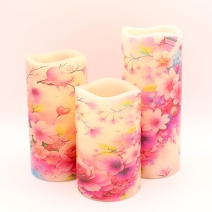 May include: Three pillar candles with a floral design. The candles are white with pink, purple, and yellow flowers. The candles are different sizes.