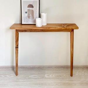 Könnte beinhalten: Eine Holzkonsolentisch mit rechteckiger Platte und vier schlanken Beinen. Der Tisch ist aus hellbraunem Holz gefertigt und hat eine natürliche Oberfläche. Zwei weiße Säulenkerzen und ein gerahmter Kunstdruck liegen auf der Tischoberfläche.
