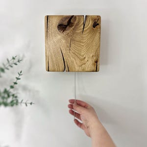 Puede incluir: Un reloj de pared cuadrado de madera con un acabado de veta natural. El reloj tiene un nudo y grietas visibles en la madera. Una cadena plateada cuelga de la parte inferior. El reloj está montado en una pared blanca.
