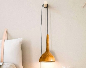 May include: A wooden pendant light with a natural wood finish hangs from a black cord. The light is turned on and illuminates the room. The light is hanging from a wooden hook on a white wall.