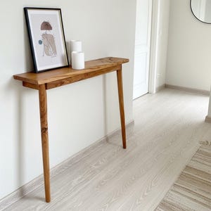 Puede incluir: Una consola de madera con acabado natural contra una pared blanca. La mesa tiene una tapa de borde vivo y patas delgadas. Una impresión artística enmarcada y dos velas blancas se exhiben en la mesa, creando una estética minimalista. La mesa mide aproximadamente 120 cm de largo.