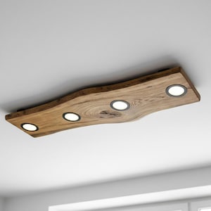 May include: A wooden ceiling light fitting with four recessed LED lights. The fixture is made from a single piece of natural-edge wood, showing the grain and texture. The lights are evenly spaced along the length of the fitting.