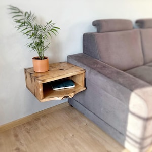 May include: A wooden wall-mounted shelf with a curved edge and a small shelf. A potted plant sits on the top shelf and a book is on the lower shelf. The shelf is mounted next to a gray couch.