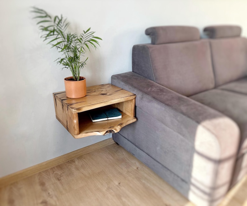 May include: A wooden wall-mounted shelf with a curved edge and a small shelf. A potted plant sits on the top shelf and a book is on the lower shelf. The shelf is mounted next to a gray couch.