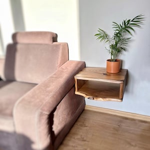 May include: A wooden wall-mounted shelf with a natural wood finish and a single shelf. A potted plant with green leaves is on the shelf. The shelf is mounted next to a light brown sofa.