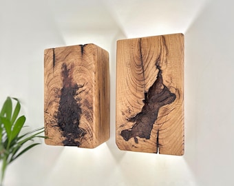 May include: Two wooden wall sconces with a natural wood grain finish. The sconces have a rectangular shape with dark, organic-shaped inlays. The wood is light brown with dark brown and black accents. The sconces are mounted on a white wall.