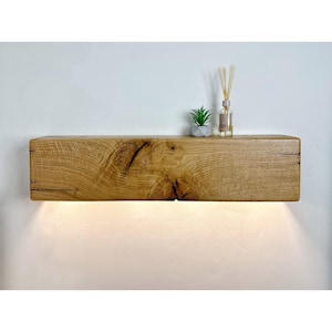 May include: A solid oak floating shelf with a natural wood grain. The shelf features integrated lighting, casting a warm glow downwards. A small potted plant and a reed diffuser are placed on the shelf, enhancing the decor.