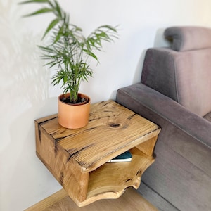 May include: A rustic wooden floating shelf with a lower shelf, mounted on a white wall next to a gray couch. A potted plant sits on the top shelf.