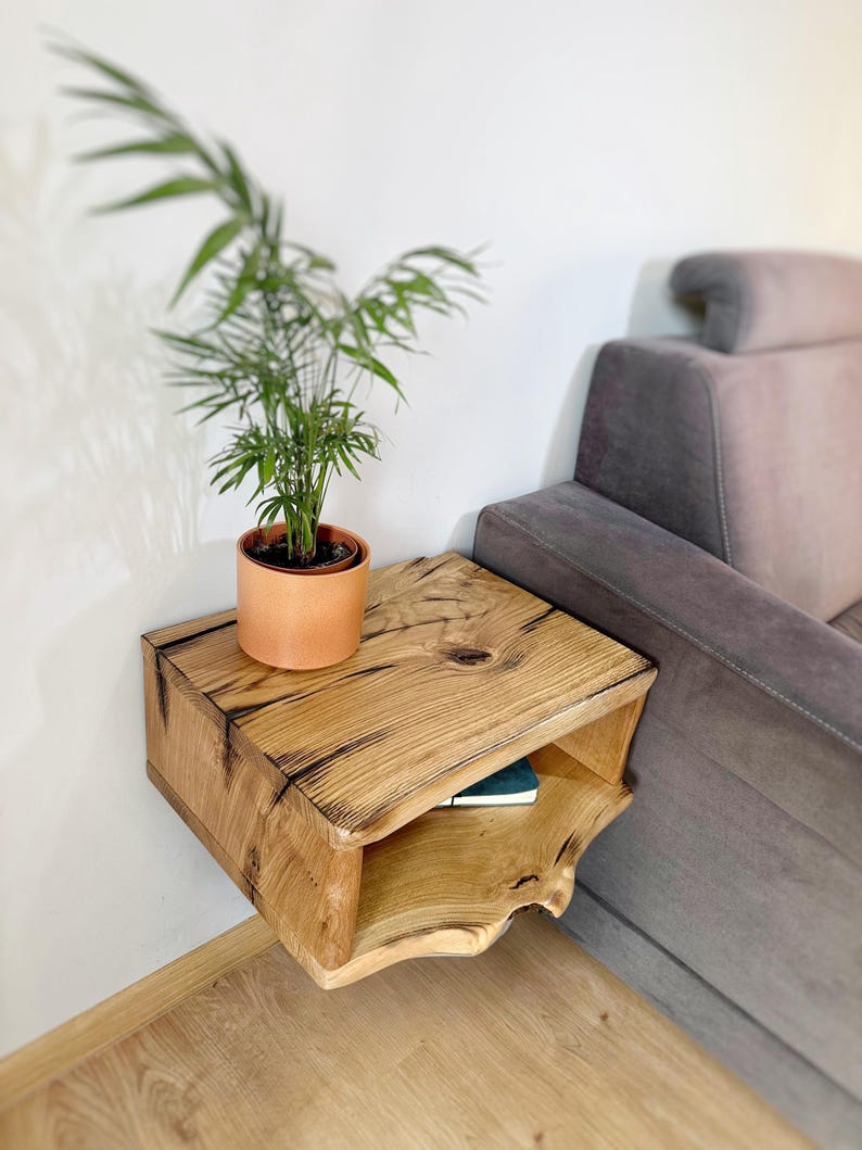 May include: A rustic wooden floating shelf with a lower shelf, mounted on a white wall next to a gray couch. A potted plant sits on the top shelf.