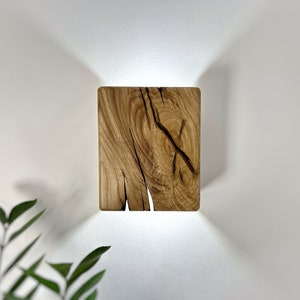 Puede incluir: Una lámpara de pared cuadrada de madera con un acabado de madera natural y una fuente de luz que brilla a través de las grietas de la madera.