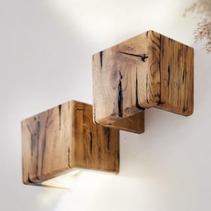Puede incluir: Dos apliques de pared de madera en forma de cubo con un acabado de veta de madera natural están montados en una pared blanca. Los apliques tienen un brillo cálido y acogedor, y la veta de la madera añade un toque rústico al diseño moderno. Los apliques son una luminaria decorativa.