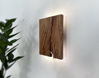 May include: A square wooden wall sconce with a natural wood finish and a light source behind it. The wood has a unique grain pattern and a knot in the center.