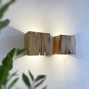 May include: Two wooden wall sconces with a light brown finish. The sconces are square shaped and have a rustic look. They are mounted on a white wall.