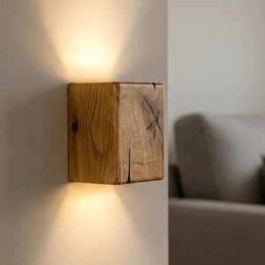 Può includere: Applique da parete in legno di forma quadrata, montata su una parete chiara. L'applique è realizzata in legno naturale con venature e nodi visibili. Emette una luce calda rivolta verso il basso, creando un'atmosfera accogliente. Il design è minimalista e rustico.
