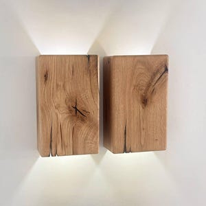 May include: Two rectangular wooden wall sconces with a natural wood grain finish. Each sconce emits a downward-facing light, creating a warm glow. The sconces are mounted on a white wall, showcasing the wood's texture and color.