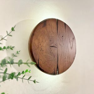 Könnte beinhalten: Runde Wanddekoration aus Holz mit natürlicher Holzmaserung. Das kreisförmige Design besteht aus mehreren Holzbrettern, die zusammengefügt wurden, wodurch eine rustikale Ästhetik entsteht. Das Holz hat eine warme braune Farbe und das Stück ist an einer weißen Wand befestigt.